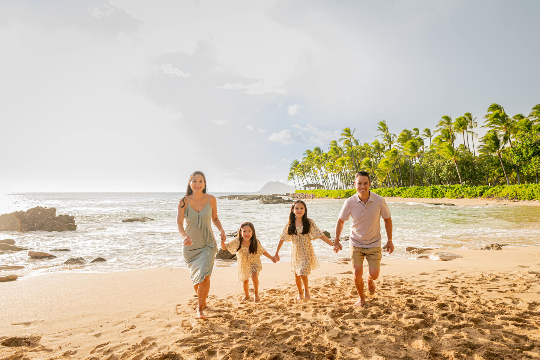 Man holding infant child up in the air while mom is laughing and smiling while they stand in the sandy shoreline on a family trip in Maui, USA